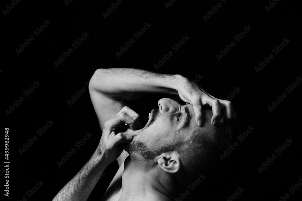 A man screams in madness clutching his face with his hands on a black background. Nervous breakdown. A clear display of emotion. - obrazy, fototapety, plakaty 