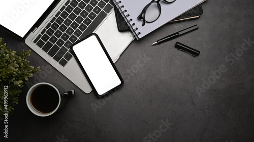 Mobile phone with blank screen  laptop computer  coffee cup and eyeglasses on dark stone background. Top view.