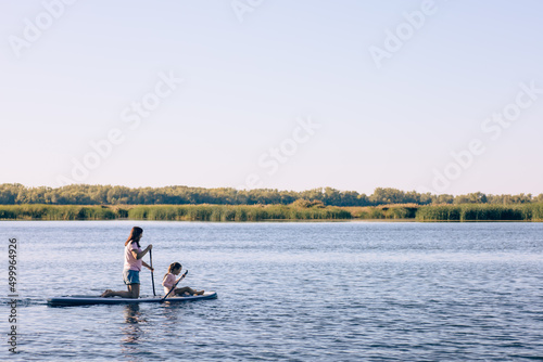 Mother and small daughter paddle boarding together on one sup on lake rowing with oars with reeds and blue sky in background. Active lifestyle. Teaching children to love sports from childhood.