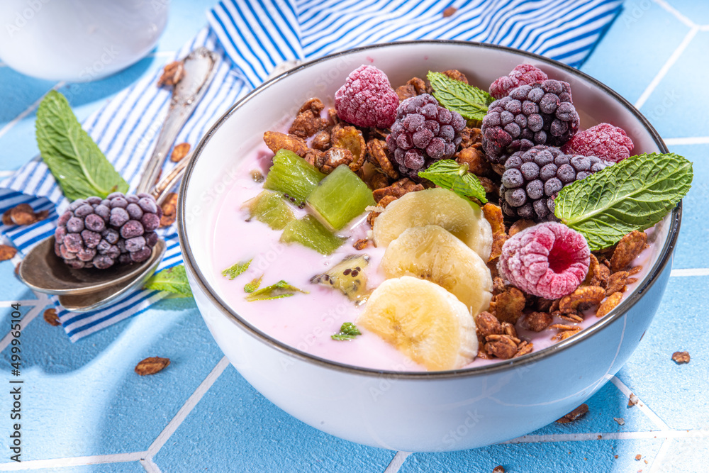 Summer breakfast healthy diet bowl with yogurt, granola, fresh fruit and berries, One portion with overnight fruity granola oats on sun-lighted tiled blue background copy space