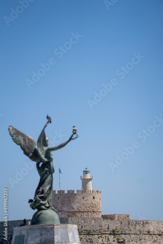 Saint Nicholas Fortress in Mandraki Harbor in focus and in the front Angel Statue of Victory .