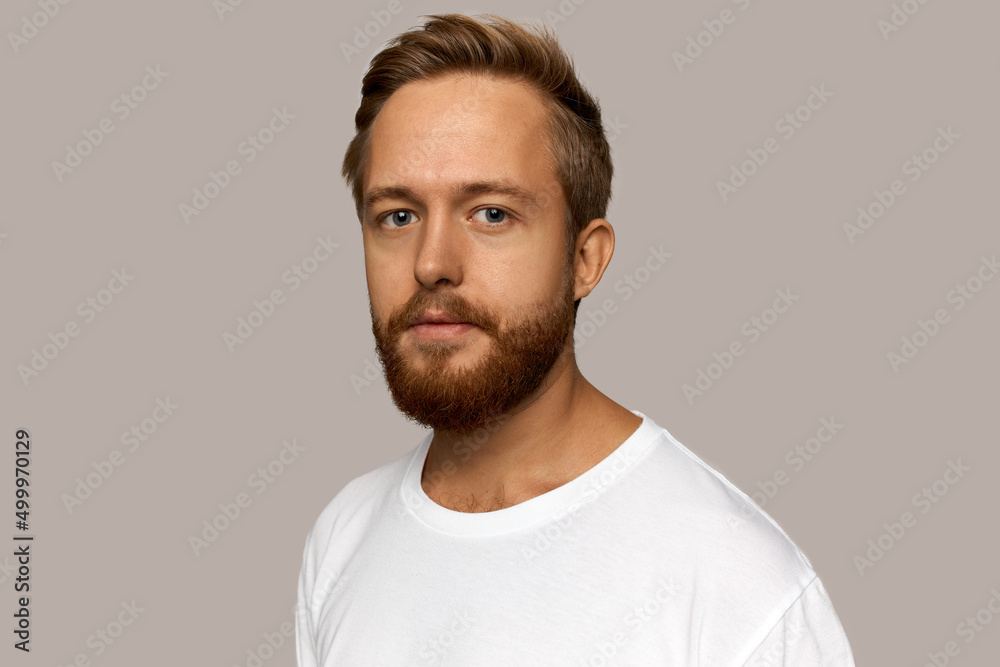 Portrait of young handsome red-haired Caucasian male hipster of 30s in white t-shirt isolated over grey background with blank copy space for your advertising content, looking positive and happy