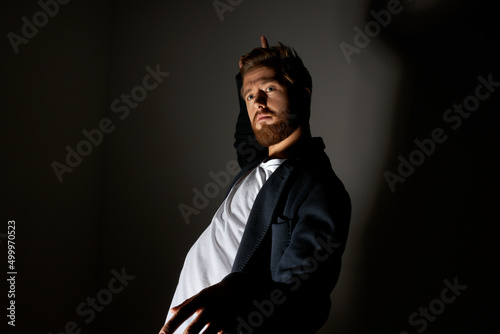 Male fashion. Portrait of charismatic photogenic ginger-headed man with beard and moustache posing in black trendy coat at studio against dark background casting shadow on wall. Light and shadow