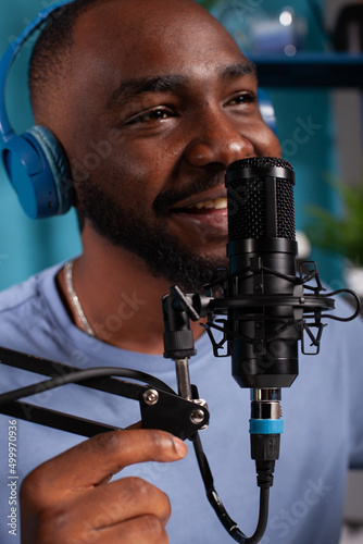 Closeup of smiling vlogger talking to audience using professional microphone wearing wireless headphones in live podcast. Close focus on studio mic used by content creator in online interview.