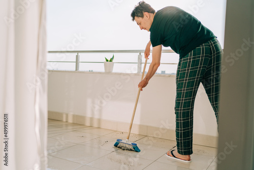 Man wiping floor with the mop. Janitor male washing the dirty floors. Guy mopping the balcony on a sunny day. Person doing home chores. Young fellow using cleaning equipment to dust the surface. photo