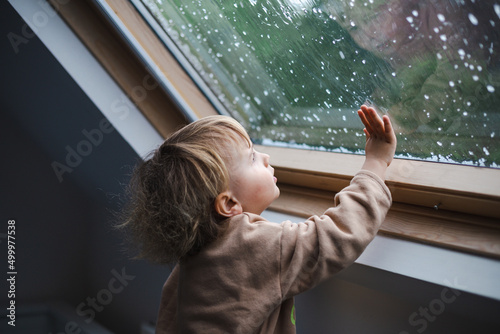 Little preschool boy staying home in bad weather and looking  with interest through window on raindrops and hail, idoors.