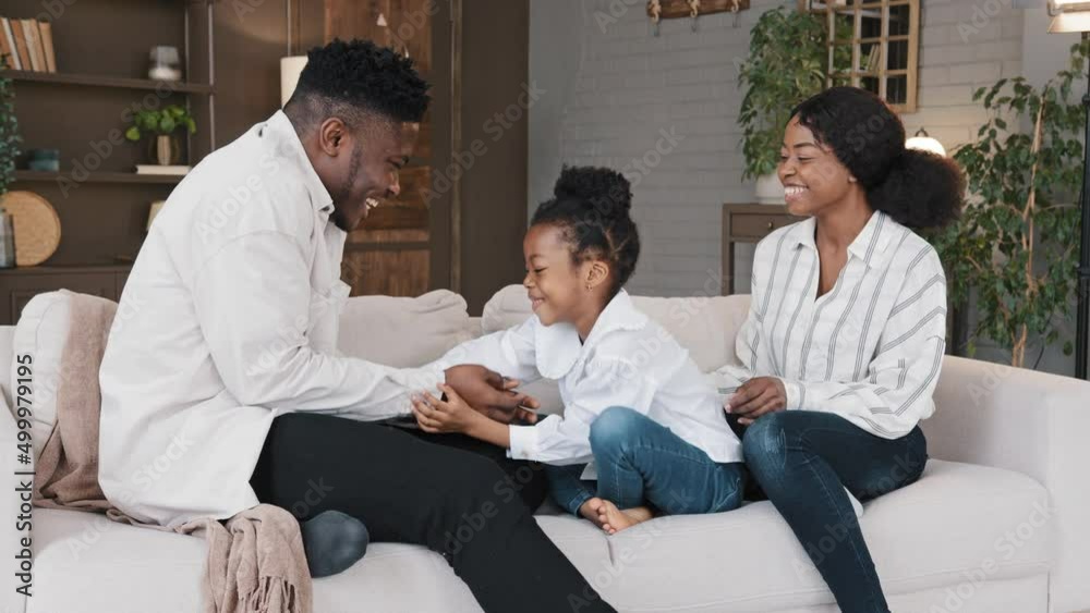 Happy african family mum dad with cute little kid daughter tickling having fun relax on sofa together laughing young carefree parents and child girl laugh play cuddling at home on couch casual game Happy african family mum dad with cute little kid daughter tickling having fun relax on sofa together laughing young carefree parents and child girl laugh play cuddling at home on couch casual game