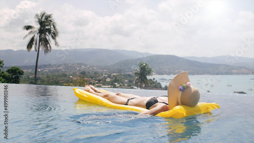 Beautiful woman in black bikini and hat swim on inflatable mattress in pool on villa