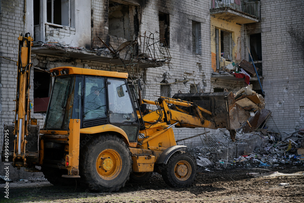 A tractor clears garbage near a destroyed town house. A large residential building without a window was damaged by an explosion during the war in Ukraine