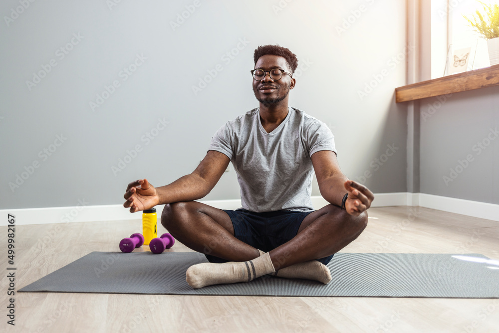 Man in sporty outfit doing yoga and meditating on an exercise mat. Sporty mindful man with tattoo meditating alone at home, peaceful calm hipster fit guy practicing yoga in lotus pose - obrazy, fototapety, plakaty 