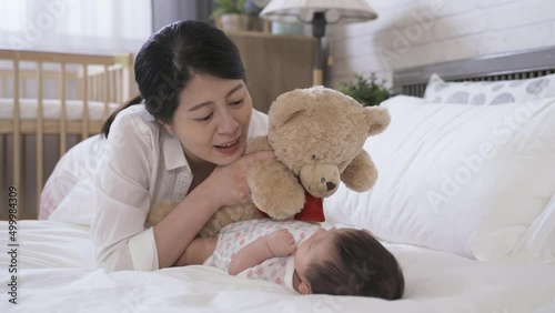 cheerful first time mother lying prone by bedside is talking to her young child with a teddy bear in hands at the bright home interior.