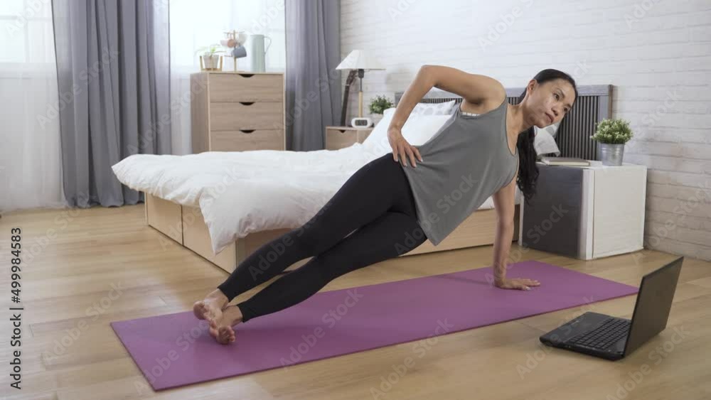 female trainer giving online training lessons at home is performing side plank by lifting her hips off the bedroom floor in front of her computer. female trainer giving online training lessons at home is performing side plank by lifting her hips off the bedroom floor in front of her computer.