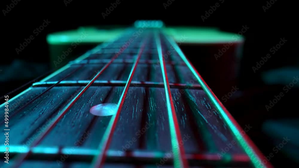 Fretboard of classical acoustic guitar against black background, illuminated by blue and red light. Brown wooden guitar neck with metal strings and frets extreme close up. Stringed musical instrument.