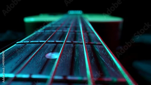 Fretboard of classical acoustic guitar against black background, illuminated by blue and red light. Brown wooden guitar neck with metal strings and frets extreme close up. Stringed musical instrument.