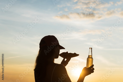 Silhouette of girl in baseball cap eating pizza and drinking soda water from glass bottle and looking at sunset sky. Rear view of female