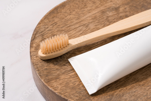 Natural bamboo toothbrush and tube with organic mineral toothpaste on wooden tray on white marble table background. White plastic tube with toothpaste. Eco bath products. Mockup