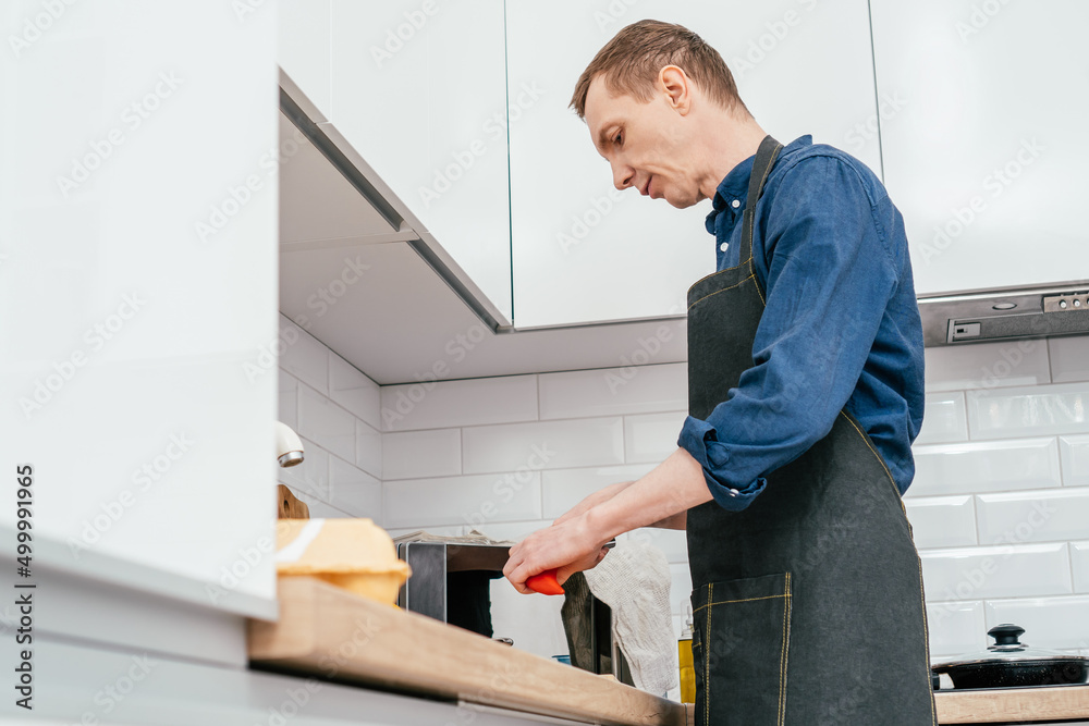 Portrait of middle-aged man wearing blue long-sleeve shirt and black apron chopping red bell pepper with knife on wooden cutting board on kitchen table. Cooking, healthy food. Portrait of middle-aged man wearing blue long-sleeve shirt and black apron chopping red bell pepper with knife on wooden cutting board on kitchen table. Cooking, healthy food.