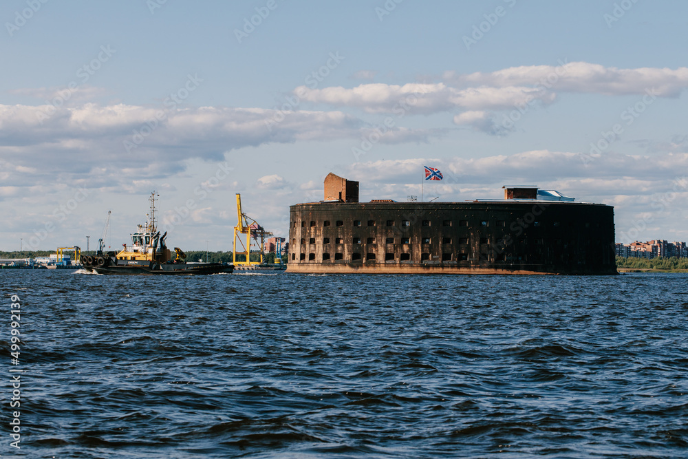 View from a boat on Fort Alexander the First in Kronstadt in the waters of the Gulf of Finland. A tugboat ship sails in the distance. Summer day