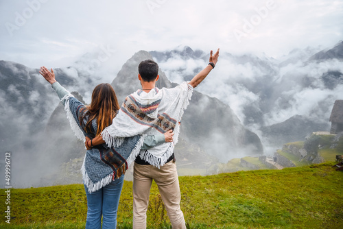 Fotografía Back view of a caucasian couple wearing the typical peruvian poncho and raising their arms in front of Machu Picchu inca city