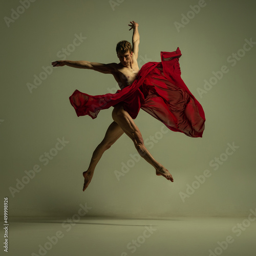 One young muscled man, flexible ballet dancer in action with red fabric, cloth isolated on olive color background. Theater, emotions, grace, art, beauty concept.