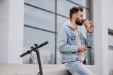 Handsome young man riding electric scooter using phone and drinking coffee