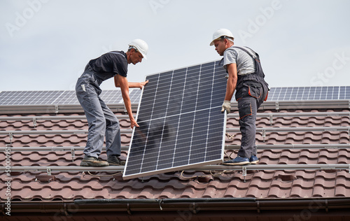 Men mounters lifting up photovoltaic solar modul on roof of house. Electricians in helmets installing solar panel system outdoors. Concept of alternative and renewable energy.