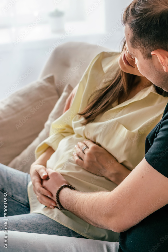 The husband hugs his pregnant wife, sitting at home on the couch..Family and pregnancy concept.Selective focus,close up. The husband hugs his pregnant wife, sitting at home on the couch..Family and pregnancy concept.Selective focus,close up.