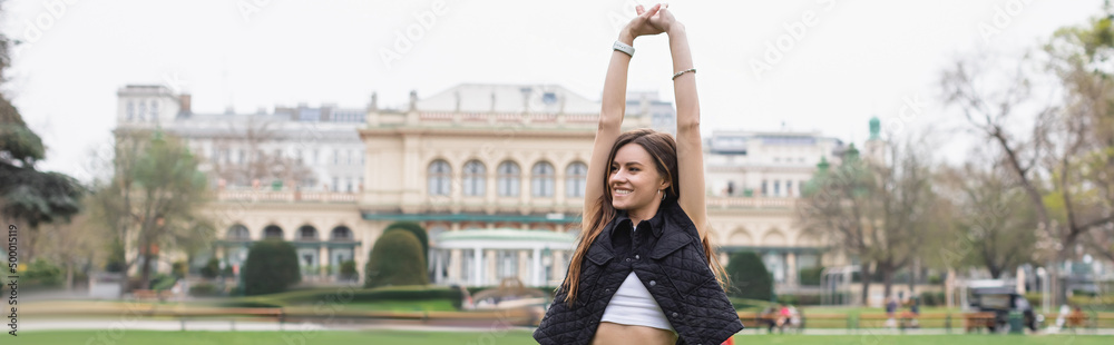 Fototapeta premium young cheerful woman in sleeveless jacket stretching in green park, banner.
