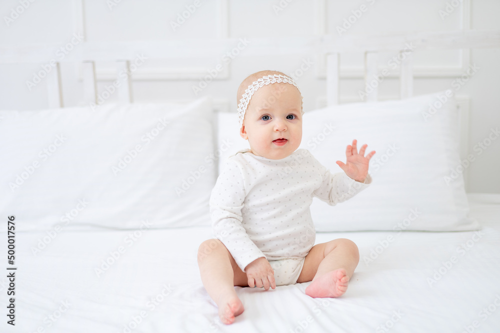 a baby girl of six months is sitting on a white cotton bed in a bodysuit on the bed at home and smiling
