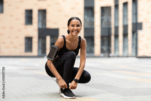 Happy young arab lady athlete in sportswear with smartphone on shoulder in headphones tying shoelaces on sneakers © Prostock-studio