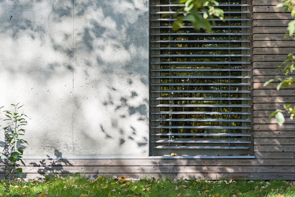 Detal architektoniczny na fragment elewacji domu jednorodzinnego wykonanego z drewna i betonu. Widoczne okno z żaluzjami aluminiowymi elewacyjnymi - obrazy, fototapety, plakaty 