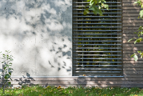 Detal architektoniczny na fragment elewacji domu jednorodzinnego wykonanego z drewna i betonu. Widoczne okno z żaluzjami aluminiowymi elewacyjnymi