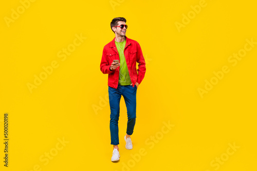Full length photo of cute brunet millennial guy go listen music look promo wear glasses shirt trousers shoes isolated on yellow background
