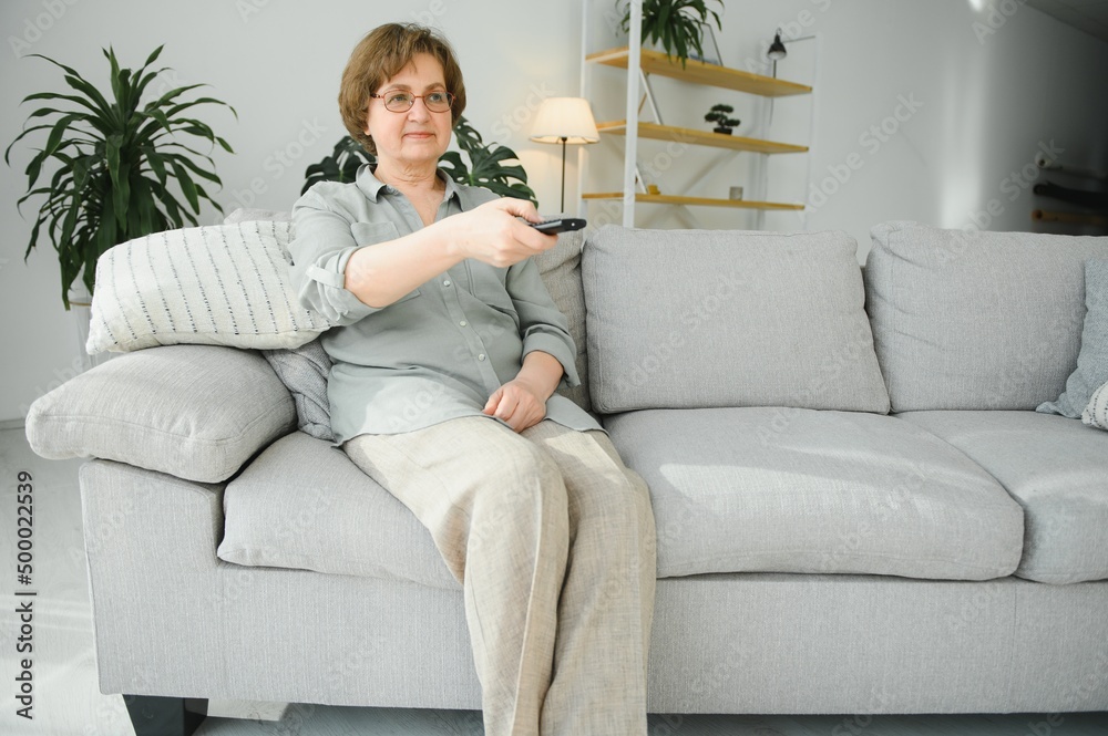 Obraz premium people, television and entertainment concept - senior woman in eyeglasses with remote control watching tv at home