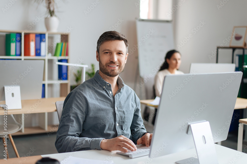 Handsome cheerful man sitting at desk, using modern computer in open space office Handsome cheerful man sitting at desk, using modern computer in open space office