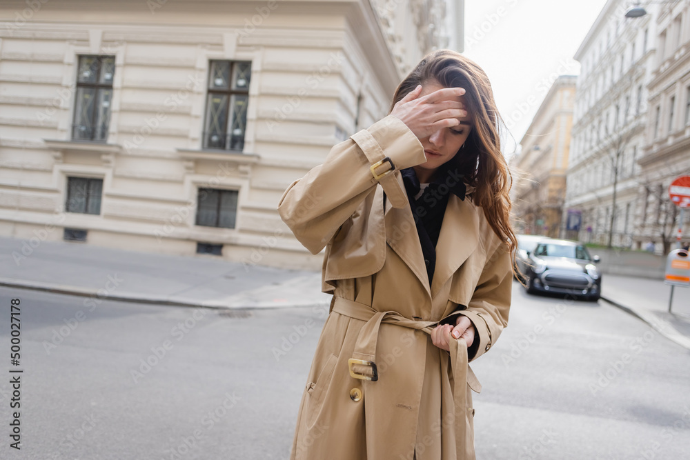 Fototapeta premium young woman in beige trench coat adjusting hair on street of european city.