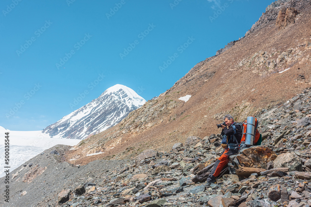 Tourist with camera sitting on stone in high mountains in sunny day. Scenic alpine landscape with man in bright sun against snow mountain peak and rocks in sunlight. Backpacker among snow mountains.