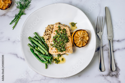 Top view of halibut fish with chimichurri sauce, green beans, lemon on a white plate on marble table
