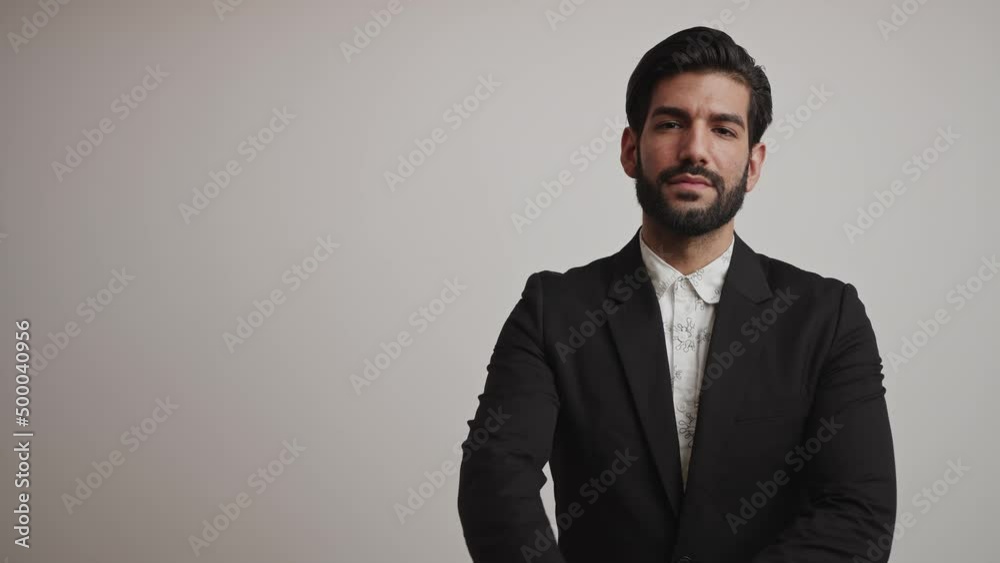 Seductive bearded Cuban businessman in a black tuxedo and white shirt crosses his arms in front of his chest and looks straight to the camera over gray background. Medium studio shot. High quality 4k