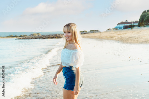 Happy smiling woman in white top and jeans shorts enjoying the moment during a walk on the seaside beach. Summer holidays, vacation. Simple pleasure. Copy space.