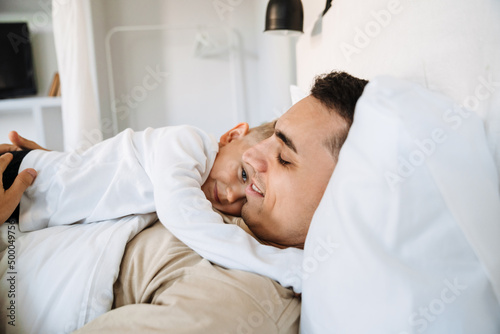 White man and his son hugging and smiling while lying on bed