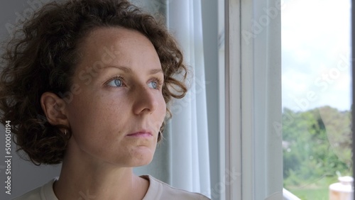Near a clean transparent window  a woman stands  looks out the window  as if waiting for changes in life  feels safe. The sad look of a woman experiencing oppression. Depression from loneliness.