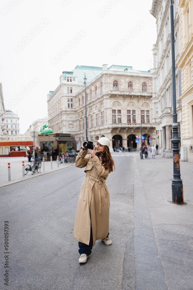 full length of young woman in stylish trench coat and baseball cap taking photo on digital camera in european city.