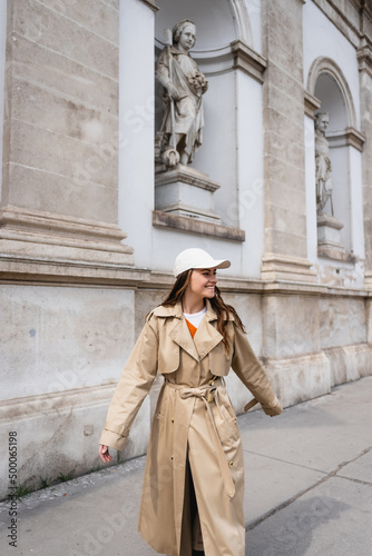 joyful young woman in stylish trench coat and baseball cap walking on street of vienna.