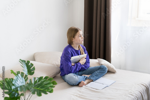 a girl in a purple hoodie is sitting on the couch with a tablet and looking away. distance education