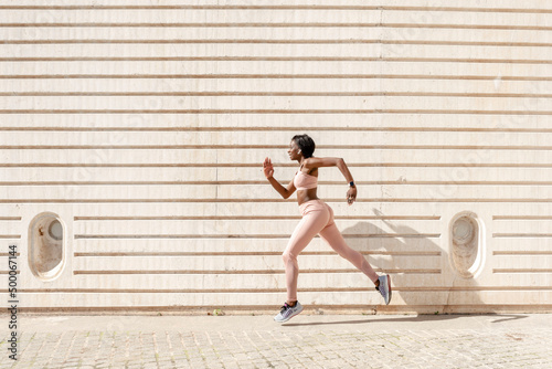 Side view of a fit young African American woman running forward against the backdrop of the city wall . Concept of sports activity and health, copy space