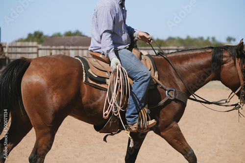 Western lifestyle shows cowboy riding bay horse through outdoor arena with rope for rodeo roping practice.
