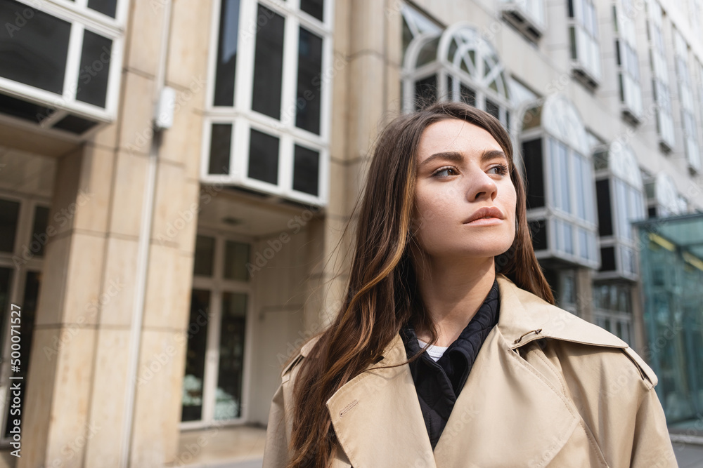 young woman in stylish trench coat looking away near building.