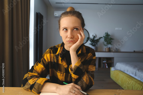young caucasian brooding woman is sitting at the desk in her bedroom at home, writing notes, rolling her eyes, making plans in her notebook, holding a pen, wearing checked yellow shirt, hair bun