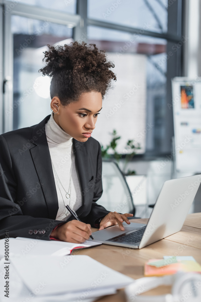 African american businesswoman using laptop and writing on notebook in office. African american businesswoman using laptop and writing on notebook in office.